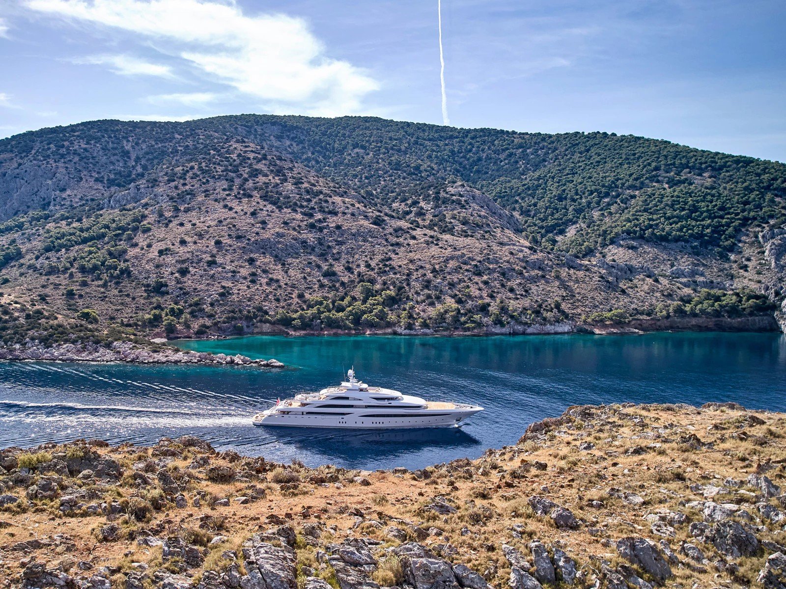 The superyacht Optasia cruising through a quiet bay below wooded hills